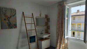 Habitación pequeña y bien iluminada con cama, mesita de noche, ventana con cortinas, estante con artículos de aseo y un cuadro de un ciervo.
