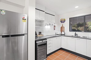A modern kitchen with a stainless steel refrigerator, white cabinets, a black countertop, and a window with blinds.