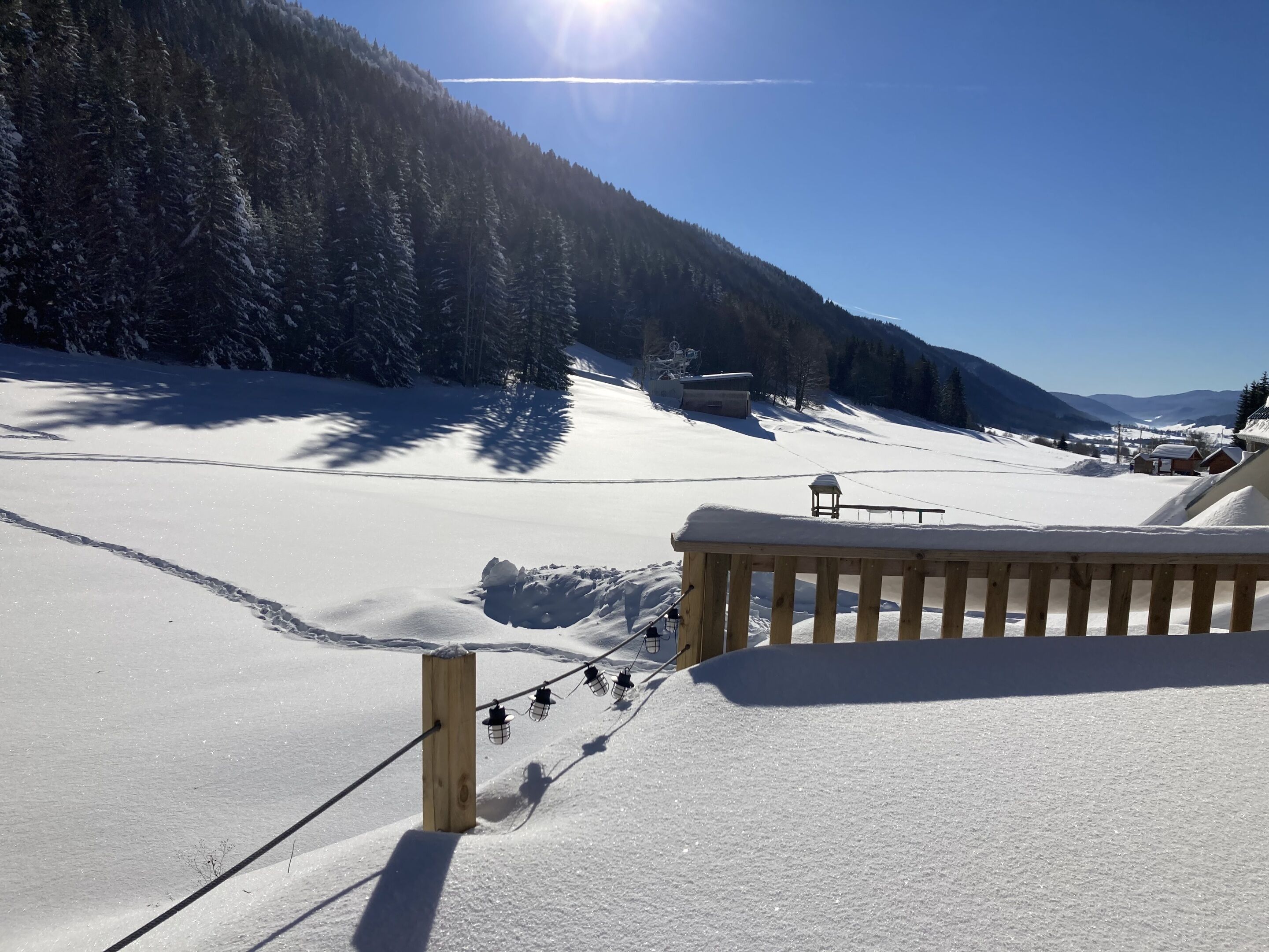 A snow-covered ski slope with a wooden railing and string lights, surrounded by forested mountains.