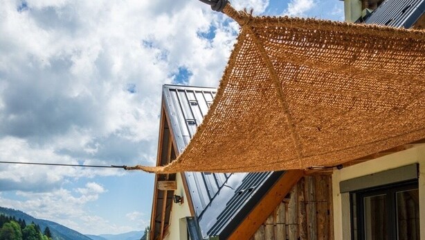 Una terrazza con tavolo e sedie, un parasole in fibra naturale e vista su un prato verde e montagne.