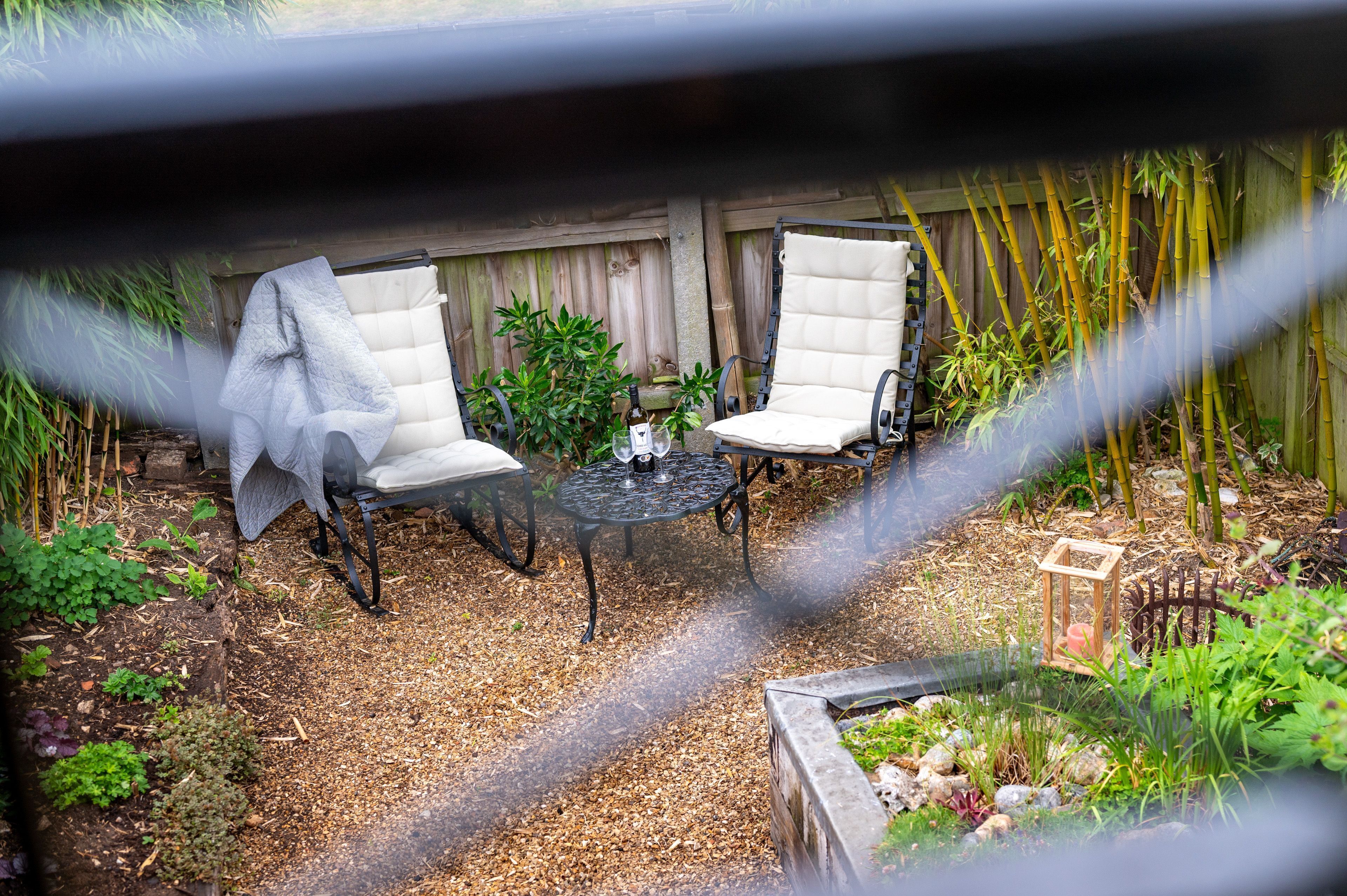 Une terrasse avec deux chaises blanches dotées de coussins et une petite table ronde, entourée de plantes en bambou et d’une clôture en bois.