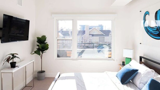 A modern bedroom with a large bed, a TV mounted on the wall, a window with a view of buildings, and a potted plant. - Art-Inspired Loft | Mountain View | Zuni Lofts (Denver)