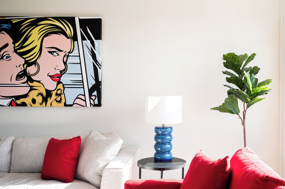 A living room with a white sofa, red cushions, a blue lamp on a small table, a green plant, and a pop art painting on the wall.