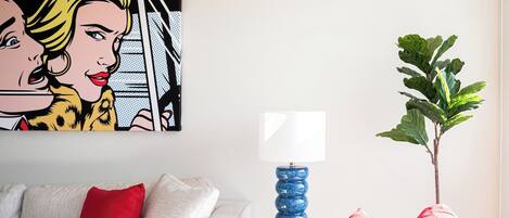 A living room with a white sofa, red cushions, a blue lamp on a small table, a green plant, and a pop art painting on the wall.