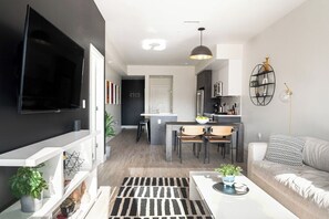 A modern living room with a flat-screen TV, a dining area with a kitchen in the background, and a sofa with patterned cushions. - Art-Inspired Loft | Mountain View | Zuni Lofts (Denver)