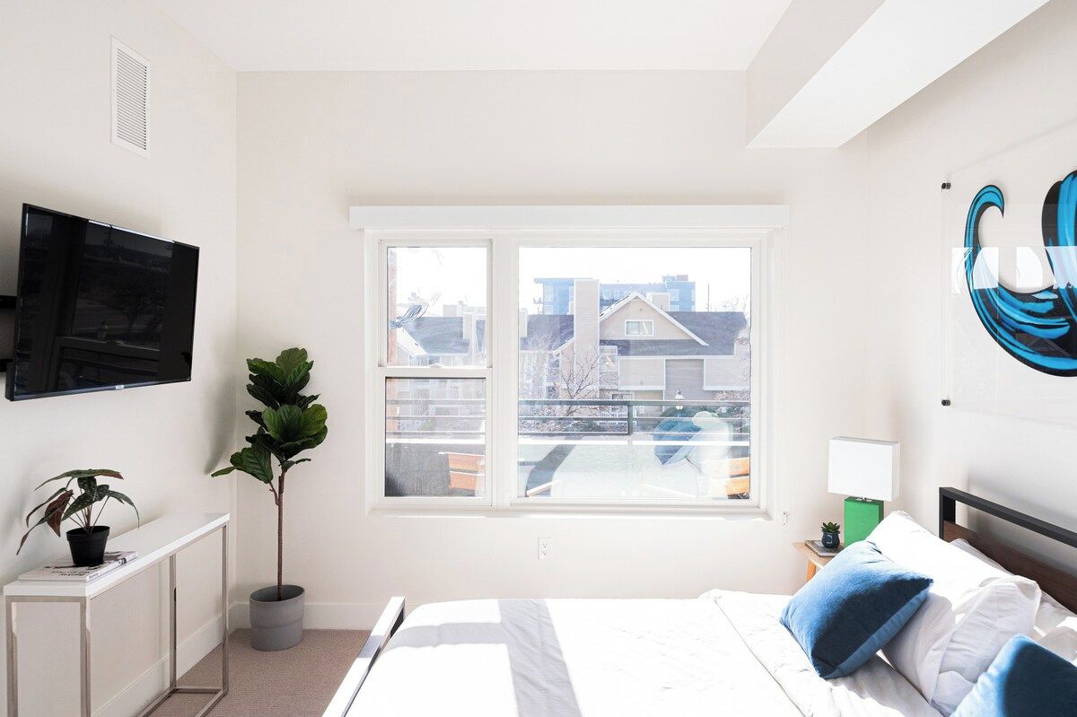 Une chambre à coucher avec un lit, un téléviseur fixé au mur, une fenêtre donnant sur des immeubles et une plante en pot.