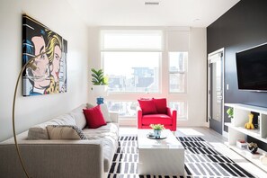 Ein modernes Wohnzimmer mit einer Couch, einem roten Sessel und einem Couchtisch. Es gibt ein großes Fenster mit Blick auf die Stadt, einen Flachbildfernseher und ein Bild an der Wand.
