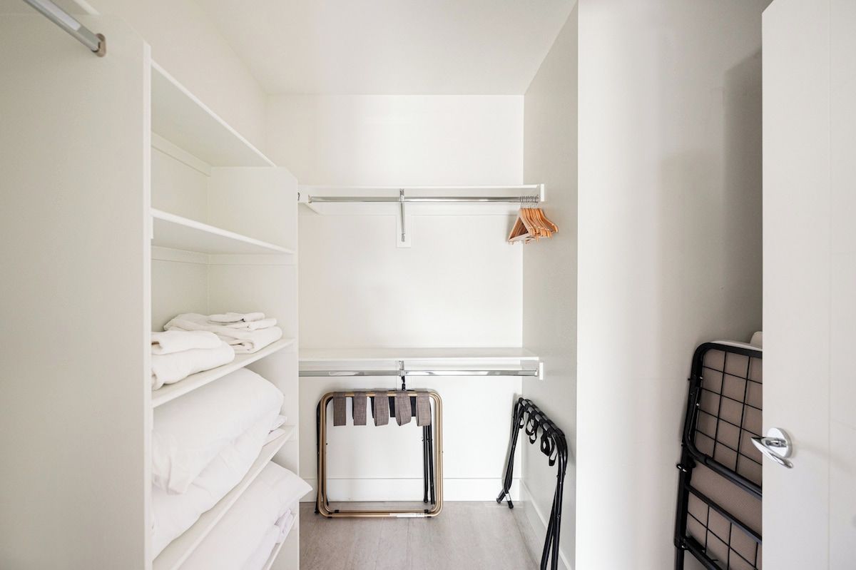 Une pièce aux murs blancs, un lit recouvert de draps blancs, un portant métallique avec des vêtements et un radiateur.