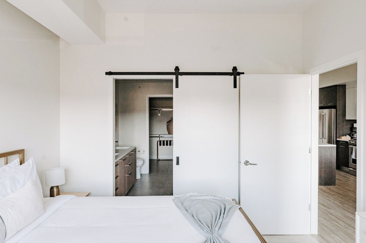 A modern bedroom with a bed, bedside table, and a view into a bathroom and kitchen area.
