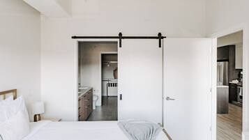 A modern bedroom with a bed, bedside table, and a view into a bathroom and kitchen area.