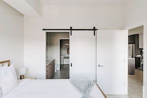 A modern bedroom with a bed, bedside table, and a view into a bathroom and kitchen area.