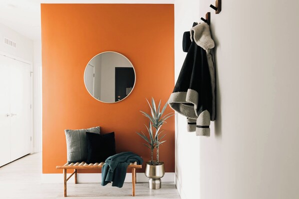 A room with an orange wall, a round mirror, a bench with cushions, a potted plant, and a coat rack with a jacket.
