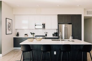 A modern kitchen with white cabinets, a stainless steel refrigerator, and a central island with bar stools. - Central LoHi Clubhouse | Skyline View | Sleeps 12! (Denver)