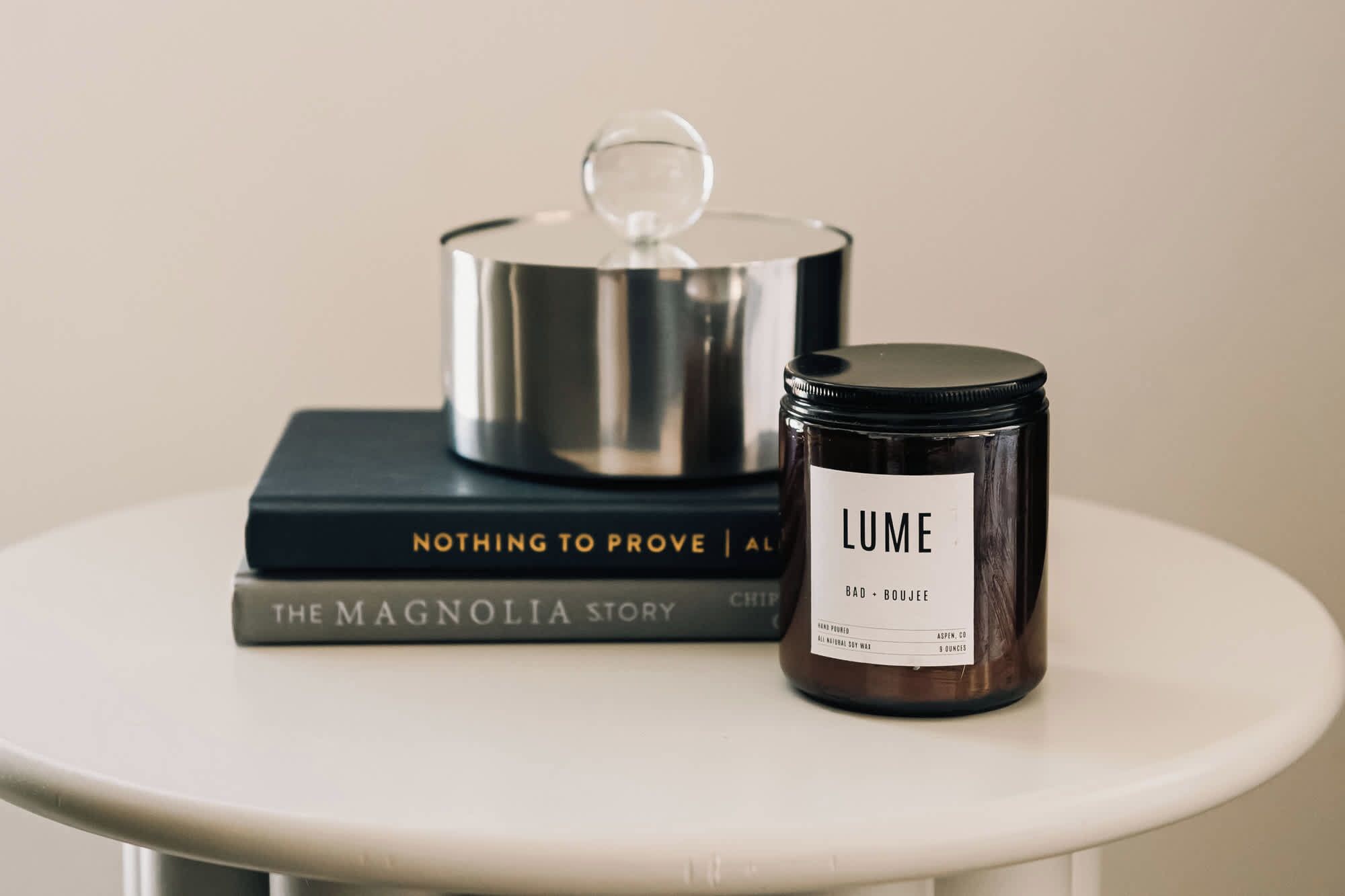 A candle labeled "LUME" on a white table with a book titled "The Magnolia Story" and a metal container with a glass lid.