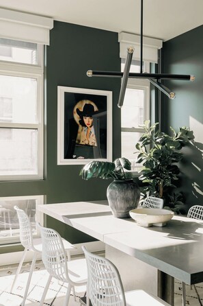 Un coin repas avec une table blanche, des chaises blanches, une grande plante en pot et un tableau encadré au mur.