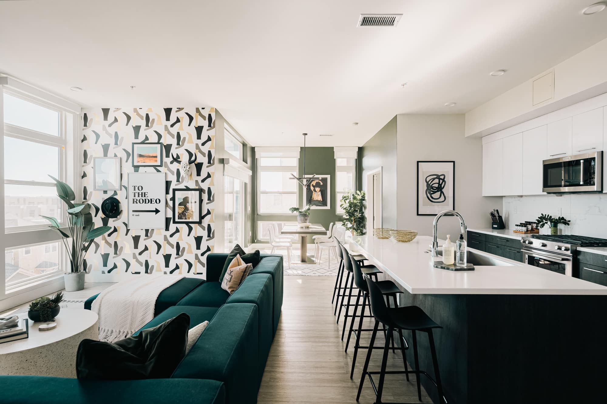 A modern kitchen with white cabinets, a central island with bar stools, and a dining area with a green sofa and a wall with framed pictures.