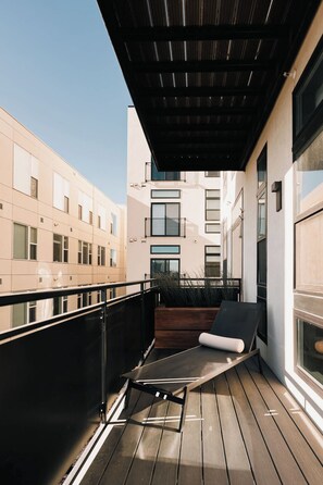 A balcony with a wooden floor, a black metal table, and a black chair. There is a wooden planter with plants and a window on the right side. - Central LoHi Clubhouse | Skyline View | Sleeps 12! (Denver)