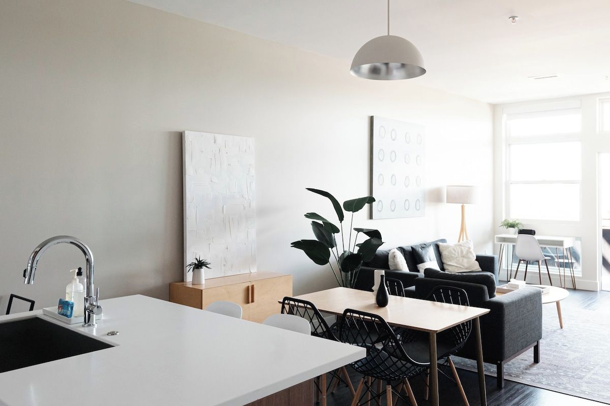 Una cocina moderna con encimera blanca, un lavamanos y una zona de comedor con mesa y sillas. Hay una planta grande y una obra de arte en la pared.