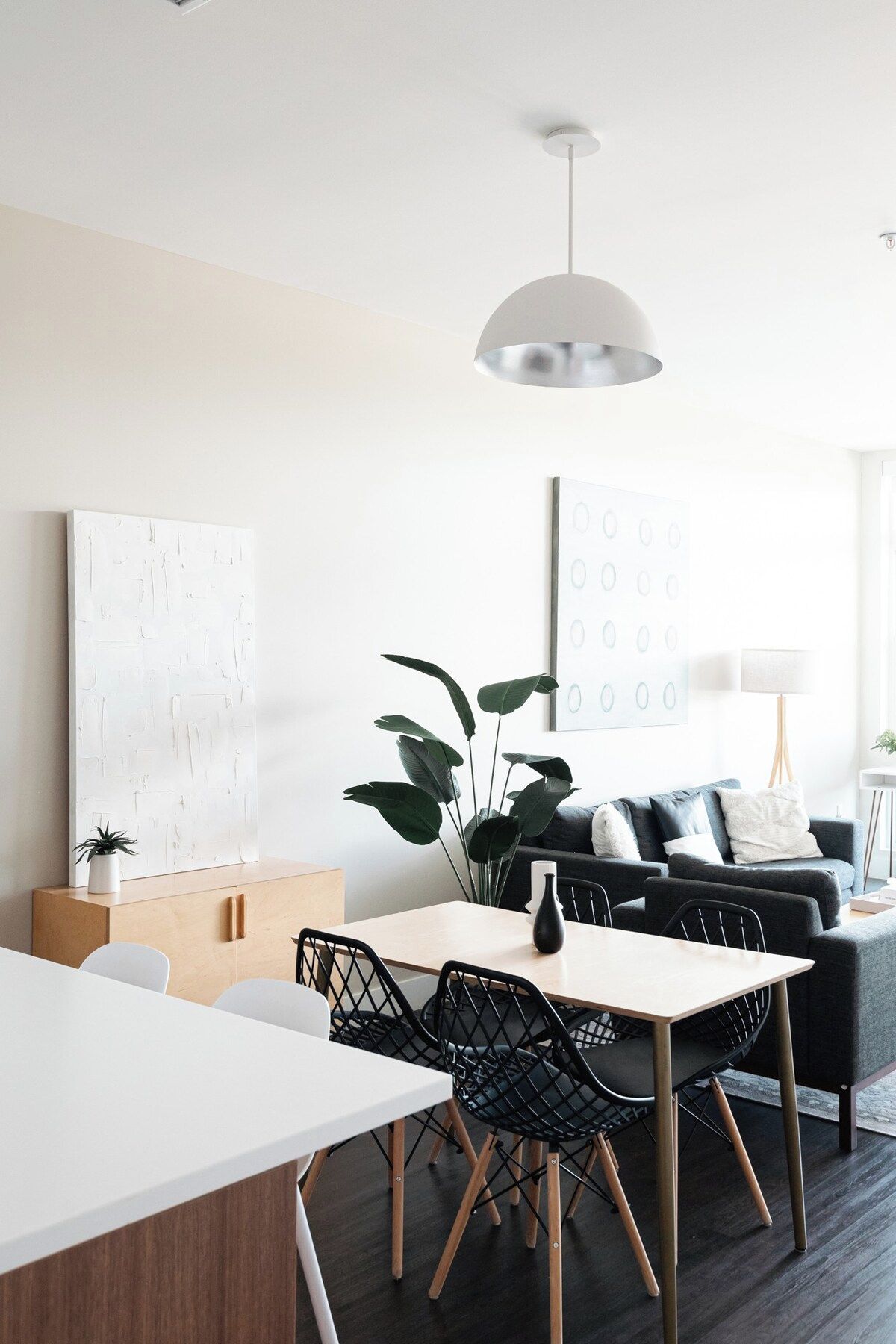 Una sala de estar moderna con comedor, que presenta una mesa blanca, sillas negras, un sofá gris y una planta grande.