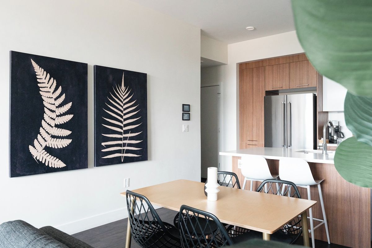 Un comedor moderno con una mesa de madera y sillas negras, una cocina con gabinetes de madera y electrodomésticos de acero inoxidable, y dos cuadros con hojas de helecho en la pared.