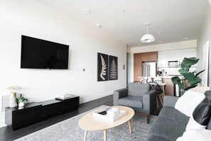 A modern living room with a flat-screen TV, grey sofas, a coffee table, and a kitchen in the background. - New Spacious Modern Loft | Center of LoHi (Denver)
