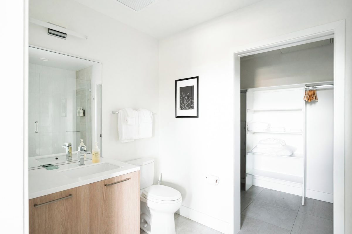 A modern bathroom with a white sink, mirror, and toilet. There is a framed picture on the wall and a towel rack with towels.