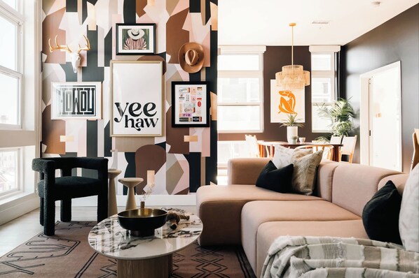 Un salon moderne avec un canapé beige, un fauteuil noir et une table basse ronde. La pièce est agrémentée d’un mur orné d’un motif géométrique, d’une grande fenêtre et d’un luminaire suspendu.