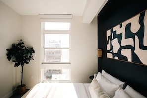 A bedroom with a large window, a potted plant, a bed with white bedding, and a wall with abstract art. - Skyline Loft | Sweeping City Views | Central LoHi (Denver)