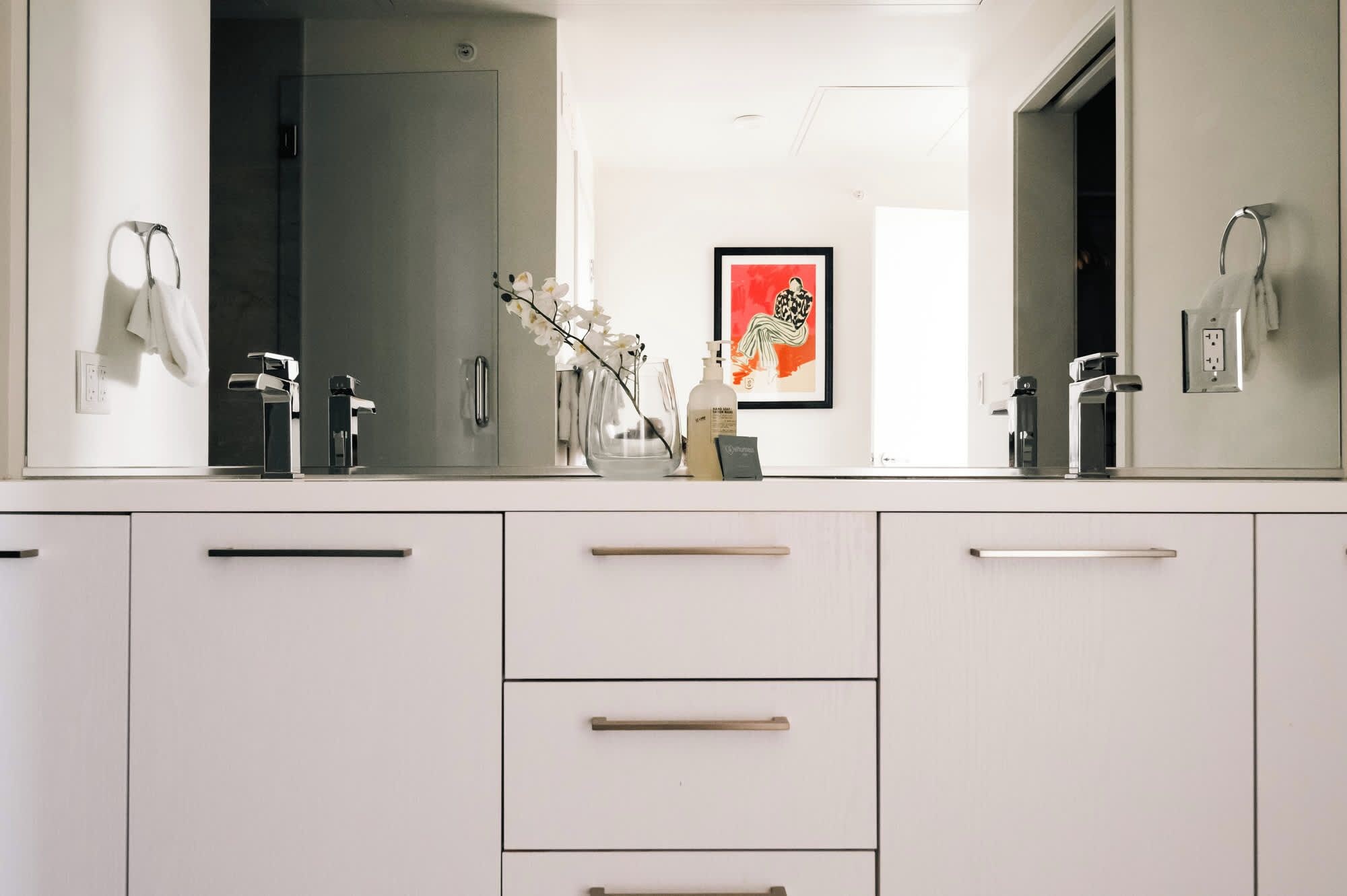 Ein modernes Badezimmer mit einem großen Spiegel, weißen Schränken und einem gerahmten Kunstwerk an der Wand.