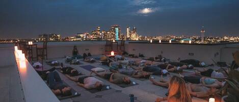 A rooftop yoga session with participants lying on mats, surrounded by candles, overlooking a city skyline at dusk.