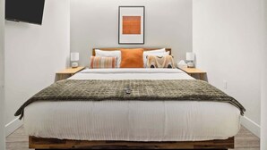 A neatly arranged bedroom with a bed, bedside tables, a framed artwork on the wall, and a television mounted on the wall. - Modern Lifestyle Loft with View | Zuni Lofts (Denver)