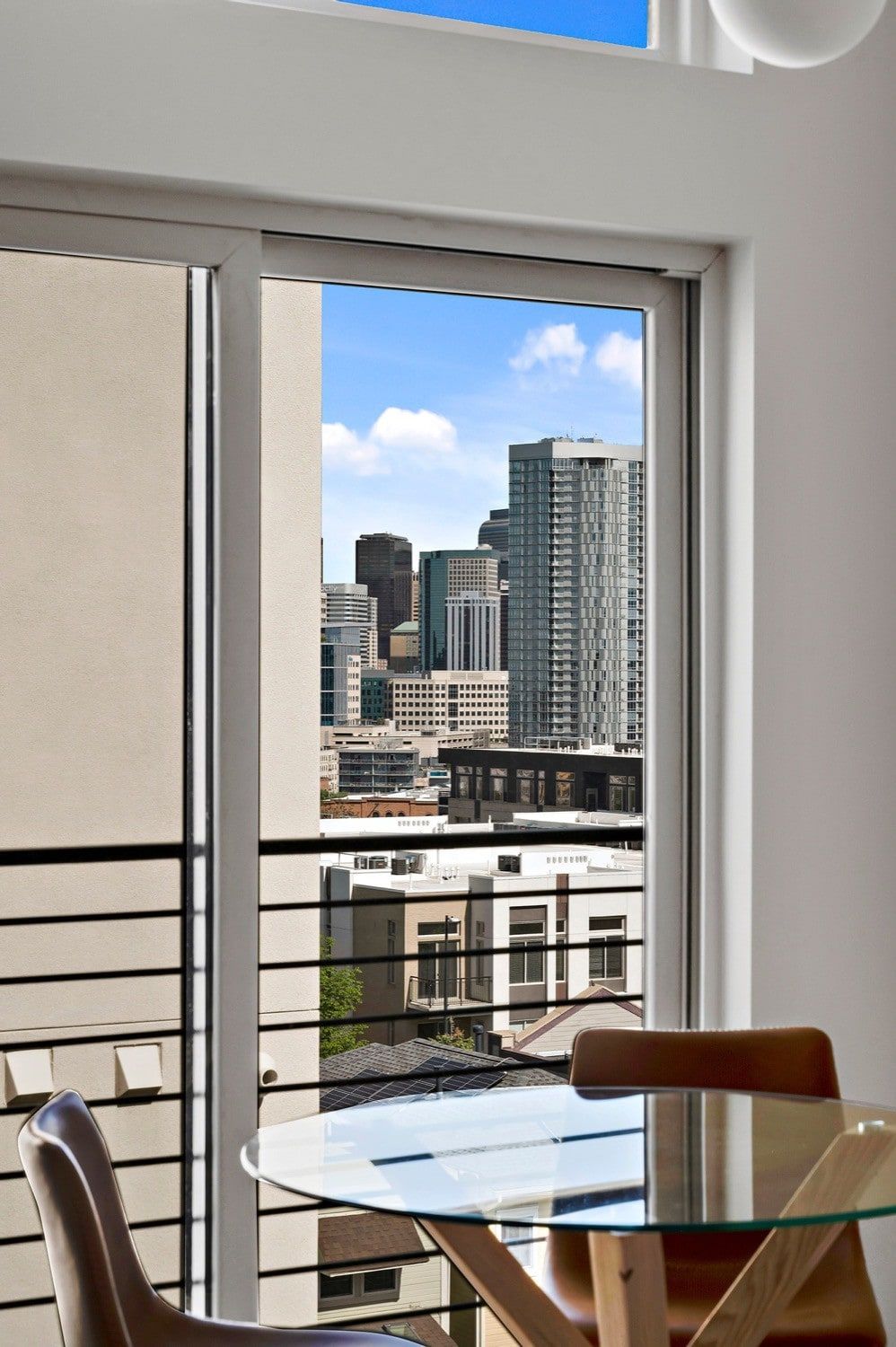 A room with a glass table and chairs, a view of a cityscape through a window, and a white wall.