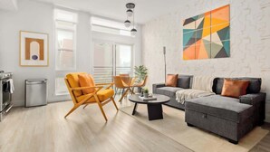 A modern living room with a grey sofa, orange armchair, and a coffee table. There is a large window providing natural light, and a geometric artwork on the wall. - Modern Lifestyle Loft with View | Zuni Lofts (Denver)