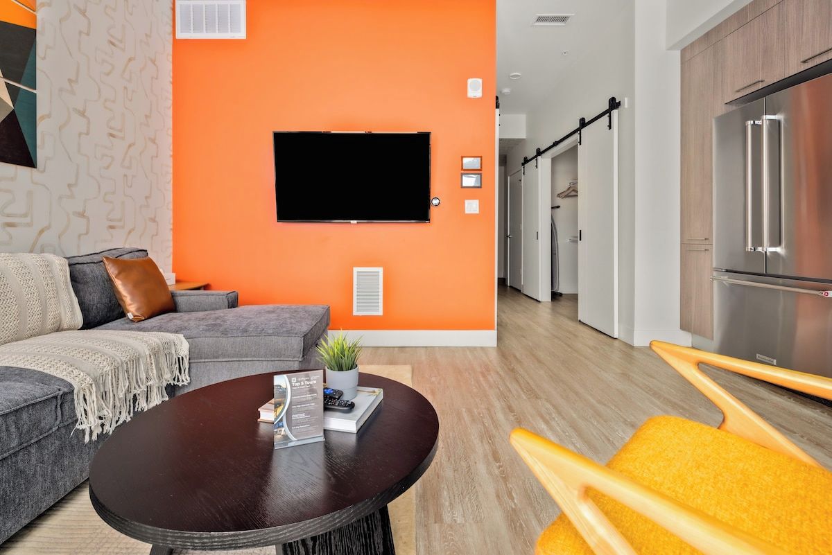 Ein modernes Wohnzimmer mit einem grauen Sofa, einem dunklen Couchtisch und einem Flachbildfernseher an einer orangefarbenen Wand.