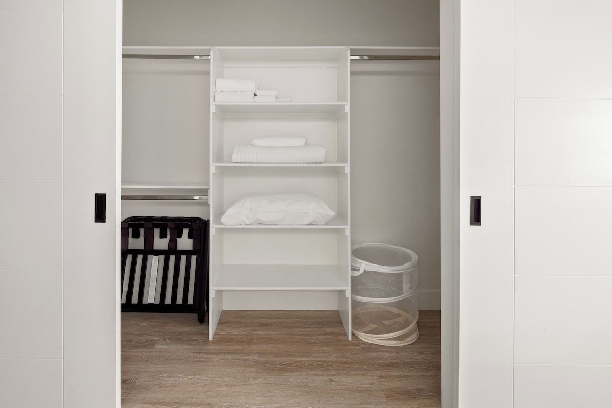 Ein weißer Kleiderschrank mit Regalen und Kleiderstange, ein schwarz-weiß gestreiftes Babybett und eine durchsichtige Kunststoff-Aufbewahrungsbox.