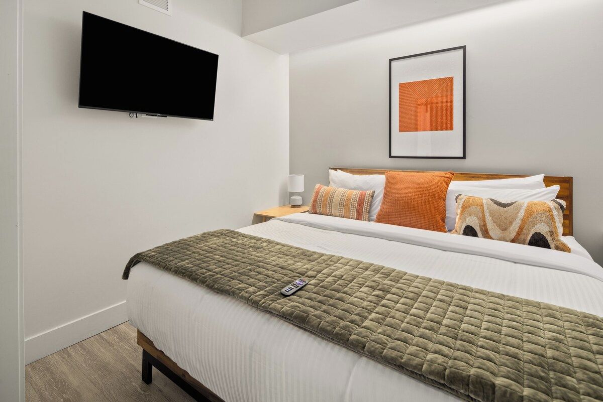 A bedroom with a bed, a flat-screen TV mounted on the wall, a bedside table with a lamp, and a framed picture on the wall.