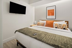 A bedroom with a bed, a flat-screen TV mounted on the wall, a bedside table with a lamp, and a framed picture on the wall.