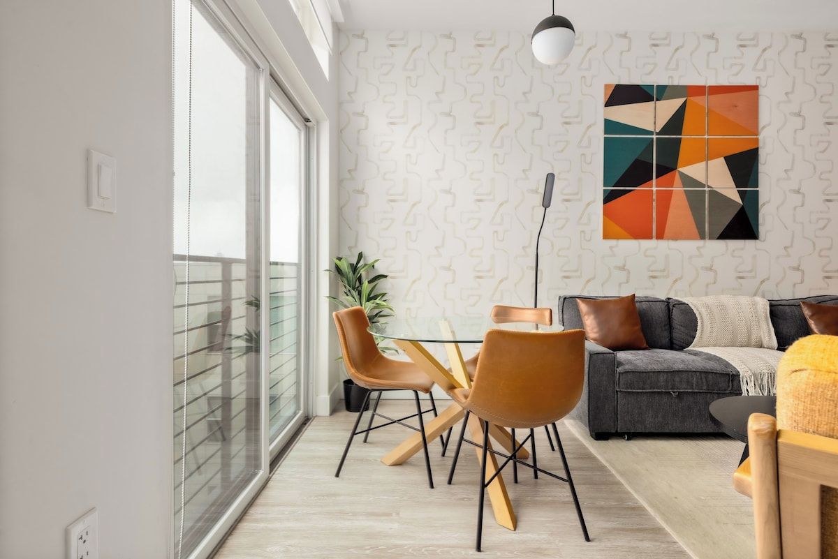 A modern living room with a grey sofa, brown leather chairs, a glass dining table, and a geometric wall art piece.