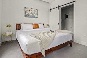 Um quarto com cama de madeira, roupa de cama branca, manta e um quadro na parede.