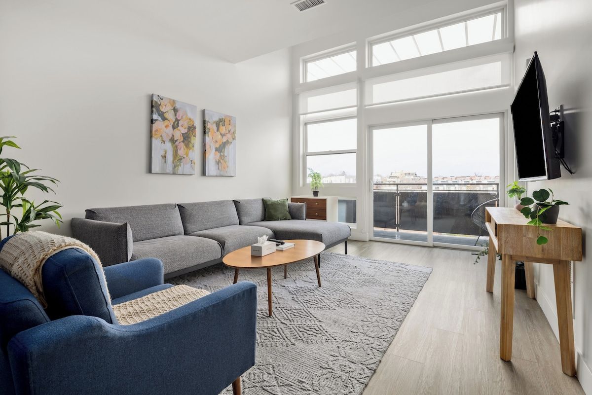 A modern living room with a grey sofa, blue armchair, and a coffee table. There are two paintings on the wall, a TV mounted on the wall, and a balcony with a view.