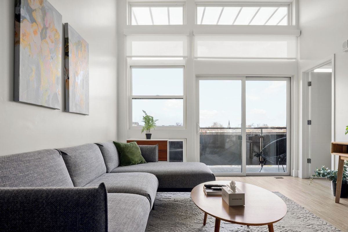 A modern living room with a grey sofa, a round coffee table, and a large window with a view of the sky and buildings.