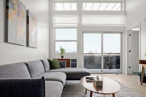 A modern living room with a grey sofa, a round coffee table, and a large window with a view of the sky and buildings.