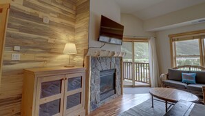 A room with a stone fireplace, wooden panel walls, a flat-screen TV, a sofa with a cushion, and a view of the outdoors. - Modern Condo with Amazing Views, Right next to the Lift - CS407 by Redawning (Copper Mountain)