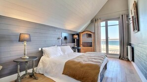 Een slaapkamer met een bed, nachtkastje, lamp, dressoir en uitzicht op het strand door een schuifdeur.