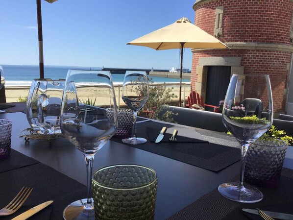 A table set with wine glasses and cutlery, overlooking a beach and a brick building.
