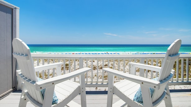 Twee witte Adirondack-stoelen op een houten terras met uitzicht op een strand, onder een helderblauwe lucht.