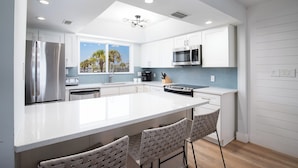 Een moderne keuken met witte kasten, roestvrijstalen apparatuur, een groot kookeiland met barkrukken, en uitzicht op palmbomen door het raam.