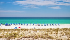Een strand met wit zand en helder turquoise water. Er zijn verschillende blauwe parasols en ligbedden.