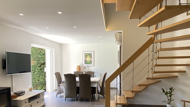 A modern living room with a flat-screen TV, a sofa, a dining area with a table and chairs, and a wooden staircase. - Villa + Studio SUR LE Golf (Saint-Raphaël)