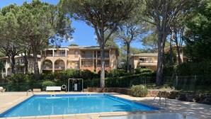 A swimming pool with a tiled deck, surrounded by trees and a building with balconies. - Villa + Studio SUR LE Golf (Saint-Raphaël)
